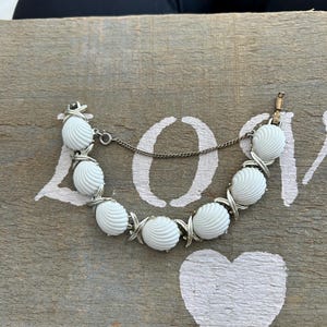 Könnte beinhalten: Ein Vintage-Armband mit weißen, muschelförmigen Perlen. Die Perlen sind durch silberfarbene Metallglieder verbunden. Das Armband liegt auf einer verwitterten Holzoberfläche mit einem gemalten Herz und dem Wort "LOVE".