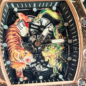 May include: A rose gold-toned tonneau-shaped wristwatch featuring a skeletonized dial with a detailed tiger and dragon illustration. The watch face includes black hour markers, central hands, and a date display window showing the number 15.