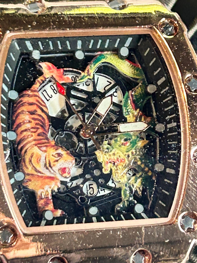 May include: A rose gold-toned tonneau-shaped wristwatch featuring a skeletonized dial with a detailed tiger and dragon illustration. The watch face includes black hour markers, central hands, and a date display window showing the number 15.