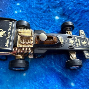 Puede incluir: Un coche de carreras de juguete negro y dorado con el texto "John Player Special". El coche tiene cuatro neumáticos negros y un motor dorado. El coche está sobre una superficie azul.