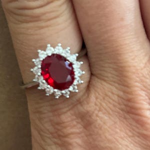 May include: A silver-toned ring featuring a large, oval-cut deep red center stone surrounded by a halo of small, clear, round-cut stones arranged in a sunburst pattern. The band is thin and metallic, supporting the prominent, multi-stone setting.