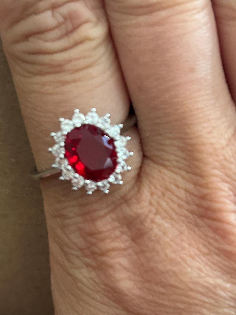 May include: A silver-toned ring featuring a large, oval-cut deep red center stone surrounded by a halo of small, clear, round-cut stones arranged in a sunburst pattern. The band is thin and metallic, supporting the prominent, multi-stone setting.