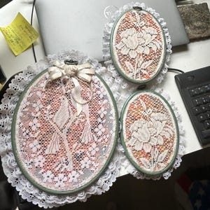 May include: Three oval-shaped lace embroidery hoops with white lace and floral designs. The hoops are framed in green and surrounded by white lace trim. One hoop has a bow.