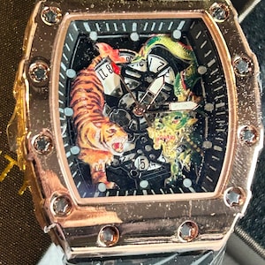May include: A rose gold-toned tonneau-shaped wristwatch featuring a skeletonized dial with a tiger and dragon graphic. The watch has a black strap, a bezel secured with eight screws, and white hour markers. The dial displays the numbers 8, 21, and 45.