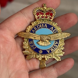 May include: A gold-colored metal badge with a crown, eagle, and text. The badge has a blue and gold circular center with the words "ARDUA AD ASTRA" and "PER" around the edge. The eagle has outstretched wings.
