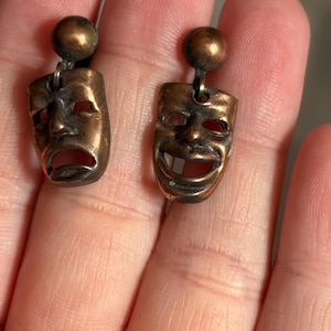 May include: A pair of bronze-toned metal earrings featuring theatrical mask pendants. One earring depicts a sad, downturned mouth mask, while the other displays a happy, smiling mouth mask. Each mask hangs from a small, round, ball-shaped stud post.