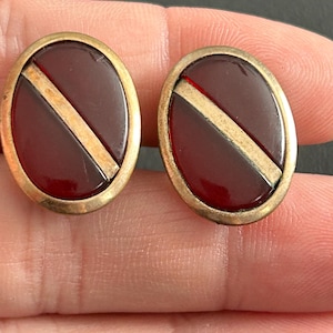 May include: A pair of vintage oval cufflinks. Each cufflink features a burgundy-colored center with a gold-colored diagonal stripe and a gold-colored border. The cufflinks are likely made of metal and glass.