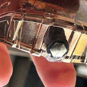 May include: A metallic gold-toned watch case with a segmented bezel and a black cylindrical crown featuring a hexagonal silver-colored end cap. The watch case is partially wrapped in clear protective plastic film, showing a reflective surface.