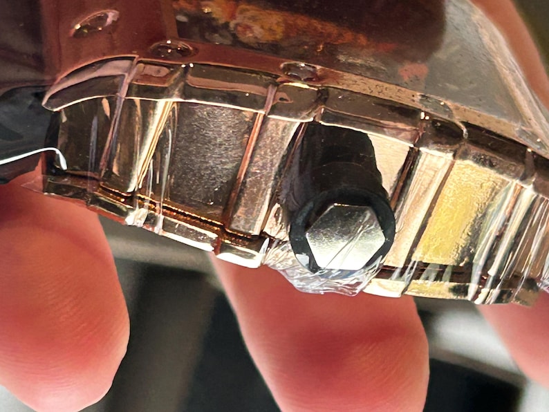 May include: A metallic gold-toned watch case with a segmented bezel and a black cylindrical crown featuring a hexagonal silver-colored end cap. The watch case is partially wrapped in clear protective plastic film, showing a reflective surface.