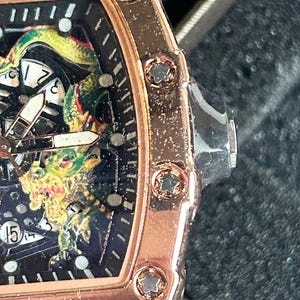 May include: A rose gold-toned watch with a tonneau-shaped case featuring star-shaped screws along the bezel. The black dial displays a detailed green and yellow dragon graphic, white hour markers, and a date window showing the number 15.