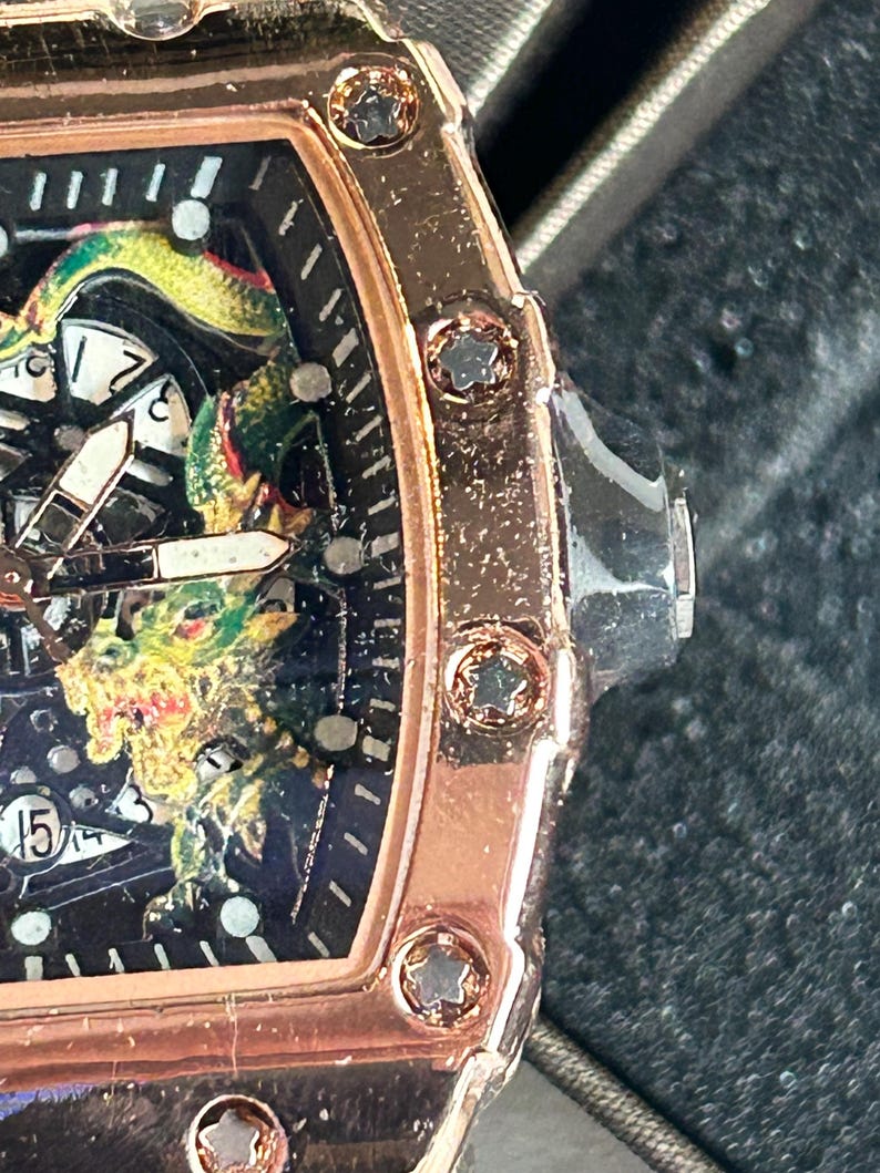 May include: A rose gold-toned watch with a tonneau-shaped case featuring star-shaped screws along the bezel. The black dial displays a detailed green and yellow dragon graphic, white hour markers, and a date window showing the number 15.