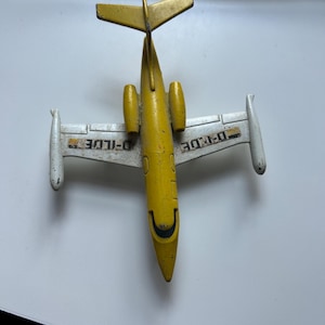 Puede incluir: Un avión de juguete vintage amarillo y blanco. El avión tiene un cuerpo, alas y cola amarillos, con puntas de ala blancas. El texto "D-ILDE" está impreso en las alas. Un juguete coleccionable clásico.