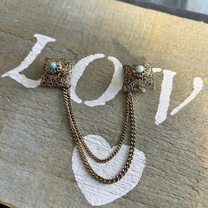 May include: A bronze-coloured vintage brooch featuring two square filigree pieces, each with a small turquoise stone. Two fine chains connect the pieces. The brooch is displayed on a wooden surface with the word "LOVE" painted on it.