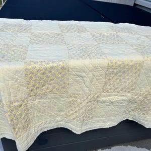 May include: A cream-coloured patchwork quilt with yellow and lavender floral squares. The quilt has a quilted texture and is made of soft fabric. The design features alternating solid cream squares.