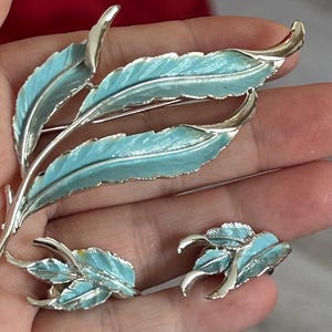 Puede incluir: Un conjunto de dos broches vintage con un delicado diseño de hojas de esmalte azul. El broche más grande tiene un diseño más elaborado con varias hojas, mientras que el broche más pequeño tiene un diseño más simple con tres hojas.