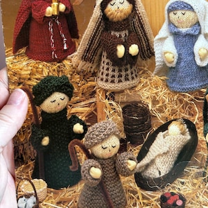 Könnte beinhalten: Gestrickte Weihnachtskrippe mit neun Figuren, darunter eine rot gekleidete Figur, eine braun gekleidete Figur, eine blau gekleidete Figur, eine grün gekleidete Figur, eine braun gekleidete Figur und eine Babyfigur in einem Korb. Die Figuren sind von Stroh umgeben.