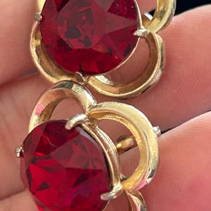 Könnte beinhalten: Ein Paar Vintage-Ohrringe in Goldoptik mit großen, facettierten, runden, rubinroten Steinen. Die Ohrringe haben ein florales Design mit einer goldfarbenen Metalleinfassung. Die Steine werden von kleinen Zinken gehalten.