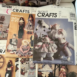 Pode incluir: Dois envelopes de moldes de costura McCall's Crafts com ilustrações de bonecas de tecido e animais de pelúcia feitos à mão. Os envelopes exibem vários designs de bonecas, incluindo gatos com roupas, com texto impresso como Fat Cats e Carole Quayle.