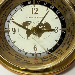 May include: A gold-toned Westclox world time watch with a cream-colored face. The watch features a world map design and displays multiple time zones. The watch has a brown leather band and a date indicator. The watch face displays the text "WESTCLOX".
