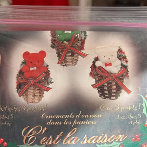 Op de afbeelding: Een knutselpakket met drie teddybeerornamenten in geweven manden, elk met een rode strik. De set bevat tekst in het Engels en Frans, met de zin "C'est la saison" in goud. Geschikt voor kinderen vanaf 5 jaar.