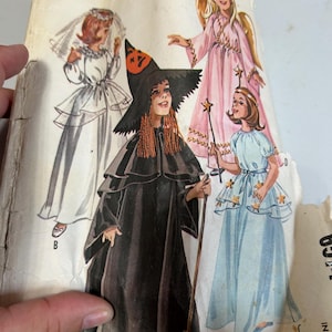 May include: Vintage costume pattern featuring illustrations of girls dressed as a witch, bride, fairy, and angel. The witch wears a black hat with an orange pumpkin, and the angel has gold wings. The text reads "Girls' Witch, Bride, Fairy and Angel Costumes."