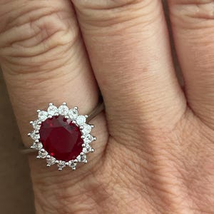 May include: A silver-toned ring featuring a large, oval-cut deep red gemstone at the center, surrounded by a halo of small, round, clear white stones set in a pronged metal frame that encircles the central red stone completely.