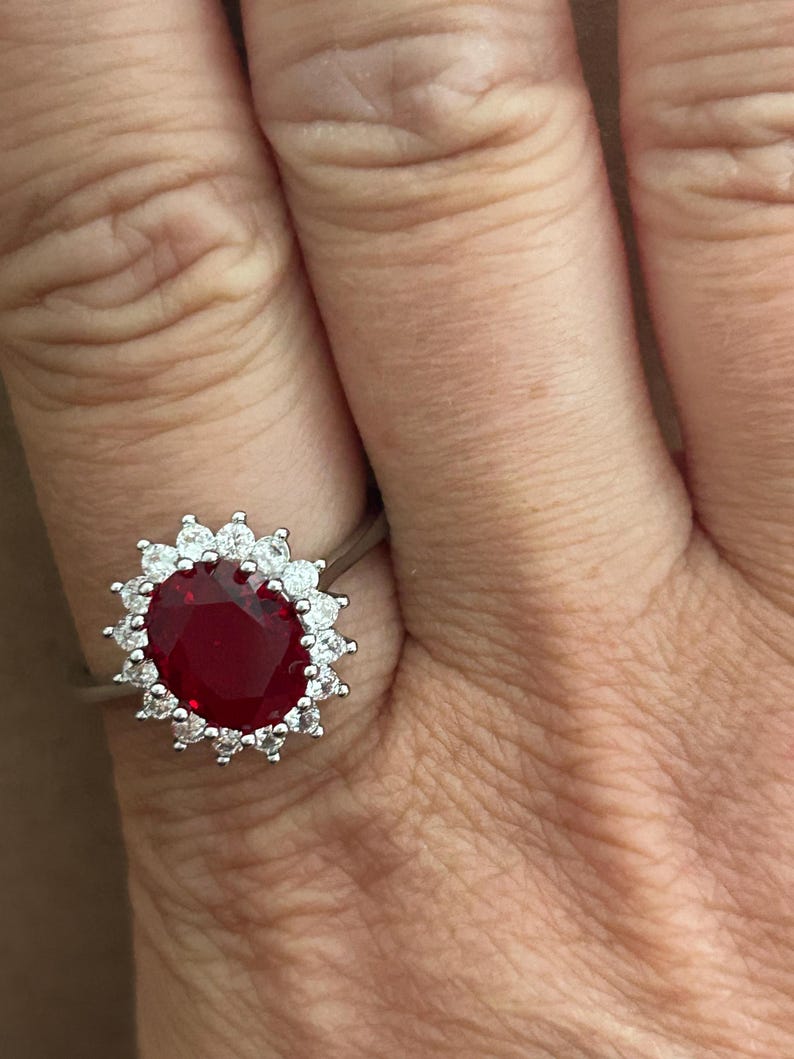 May include: A silver-toned ring featuring a large, oval-cut deep red gemstone at the center, surrounded by a halo of small, round, clear white stones set in a pronged metal frame that encircles the central red stone completely.
