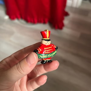 May include: A small, painted Christmas ornament in the shape of a toy soldier. The soldier wears a red uniform with gold accents and a black hat. A green banner with the words "Merry Christmas" is wrapped around the soldier.