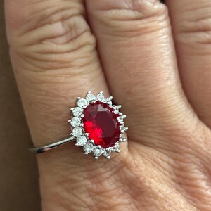 May include: A silver-toned ring featuring a large, oval-cut deep red gemstone at the center, surrounded by a halo of small, clear, round-cut stones. The band is thin and metallic, providing a contrast to the vibrant red centerpiece.