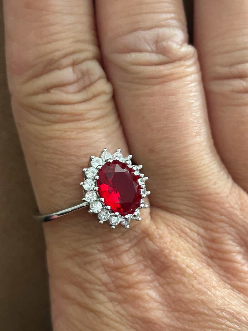 May include: A silver-toned ring featuring a large, oval-cut deep red gemstone at the center, surrounded by a halo of small, clear, round-cut stones. The band is thin and metallic, providing a contrast to the vibrant red centerpiece.