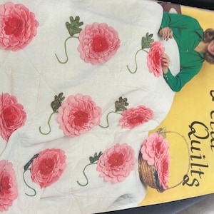 May include: A white quilt with large pink flower appliqués and green leaves. The quilt is on a yellow background with the words "Petal Quilts" in a script font. A woman in a green shirt is holding the quilt.