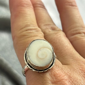 May include: A silver ring featuring a circular, off-white Shiva eye shell inlay. The shell displays a natural, light brown spiral pattern centred on its surface. The metal setting has a textured, scalloped edge surrounding the stone.