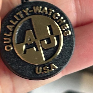 May include: A circular black tag with a gold-colored metallic center featuring the letters AJ. The text QULALITY-WATCHES is embossed along the top edge, and the letters USA are embossed at the bottom. A black cord is attached to the top of the tag.