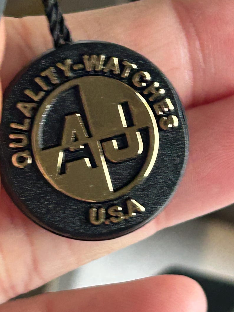 May include: A circular black tag with a gold-colored metallic center featuring the letters AJ. The text QULALITY-WATCHES is embossed along the top edge, and the letters USA are embossed at the bottom. A black cord is attached to the top of the tag.