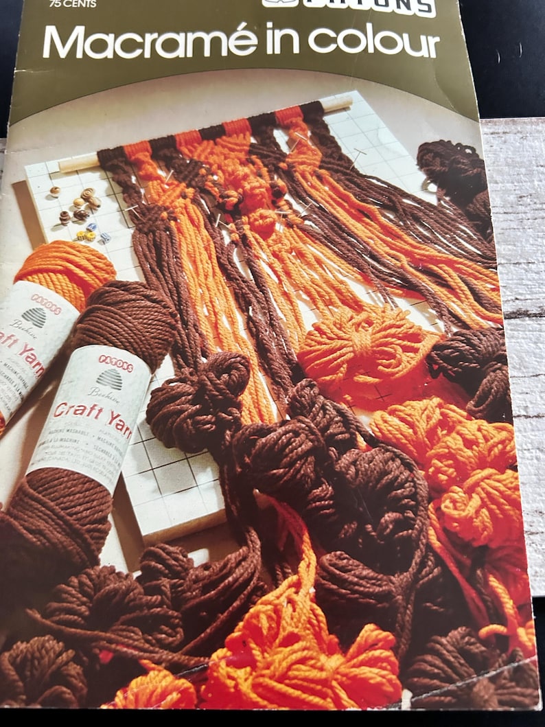 Op de afbeelding: Een vintage boekomslag getiteld "Macram&eacute; in colour" met een macram&eacute;project in uitvoering. Het project gebruikt bruin en oranje garen, met spoelen bruin en oranje handwerkgaren op de voorgrond. De omslag is groen en wit.