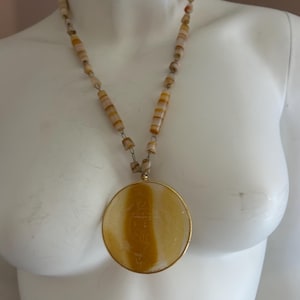 May include: A necklace with a large, round pendant and a beaded chain. The pendant has a gold-colored frame and a yellow and white striped design. The necklace is displayed on a white mannequin.