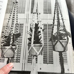 Op de afbeelding: Drie macram&eacute; plantenhangers met geweven manden, elk met een plant. De hangers vari&euml;ren in ontwerp, met kralen en kwastjes. De achtergrond toont een raam met jaloezie&euml;n. De afbeelding is gelabeld met de letters A, B en C.