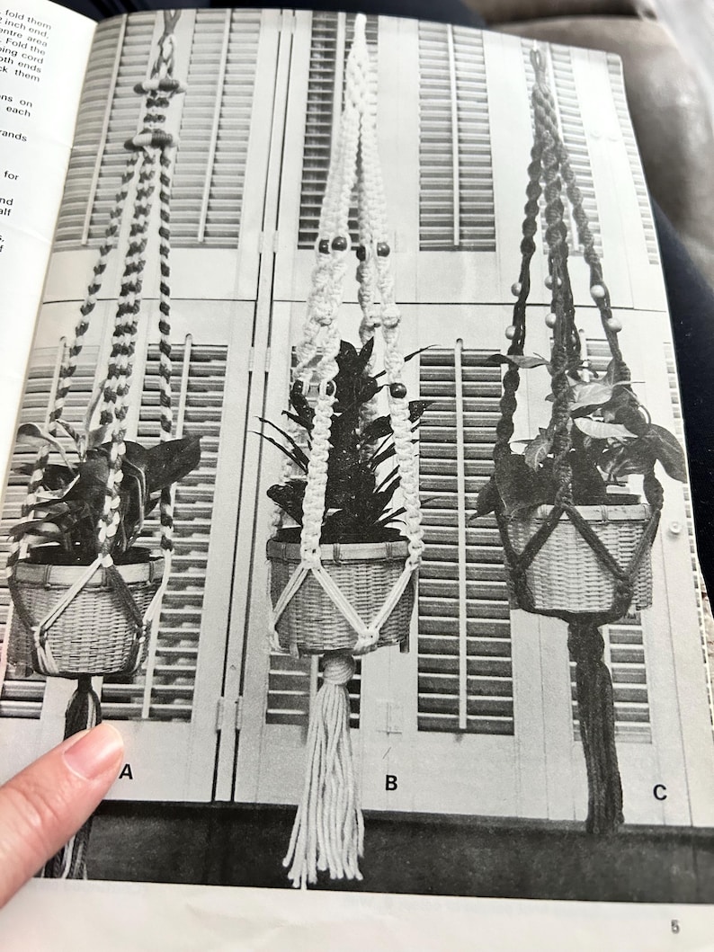 Op de afbeelding: Drie macram&eacute; plantenhangers met geweven manden, elk met een plant. De hangers vari&euml;ren in ontwerp, met kralen en kwastjes. De achtergrond toont een raam met jaloezie&euml;n. De afbeelding is gelabeld met de letters A, B en C.