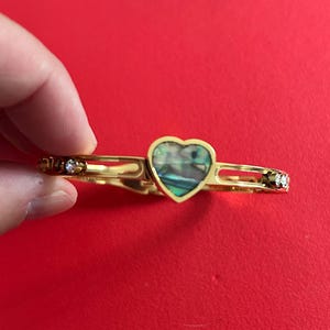 Peut inclure: Un bracelet jonc articulé doré avec un pendentif central en forme de cœur incrusté de nacre d'ormeau irisée. Le bracelet présente des découpes rectangulaires et est orné de petites pierres rondes transparentes de chaque côté.
