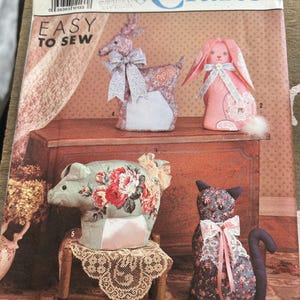 Puede incluir: Patrón de costura Simplicity Crafts 9644. El patrón incluye instrucciones para crear figuras de animales de tela, como un ciervo, un conejo, un cerdo y un gato. La portada muestra las manualidades terminadas, con telas florales.