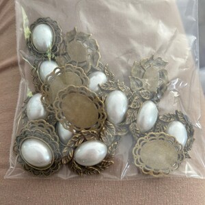 Peut inclure: Une collection de cabochons ovales avec des perles artificielles blanches lisses serties dans des cadres en métal couleur bronze antique. Certains cadres ont des bords festonnés, d'autres des motifs de feuilles, le tout dans un sac en plastique.