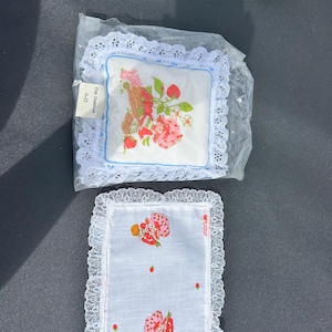 May include: Two white pin cushions with lace trim. One cushion is square with a strawberry design and a blue border. The other cushion is rectangular with a strawberry pattern. The cushions are on a dark surface.
