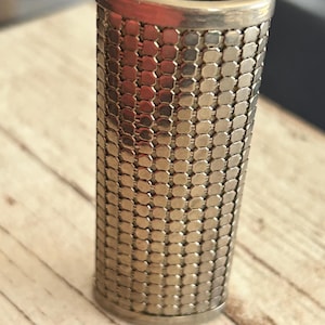 May include: A cylindrical, vintage metal mesh object with a gold-toned finish. The mesh is made of small, connected rectangular pieces. The top and bottom edges are smooth, and the interior is dark. The object is likely a decorative item or a container.