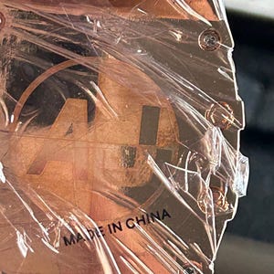 May include: A square-shaped copper-toned watch case back featuring a circular logo with the letters AU inside. The metal surface is covered in a protective clear plastic film and includes four visible screws at the corners with the text MADE IN CHINA.