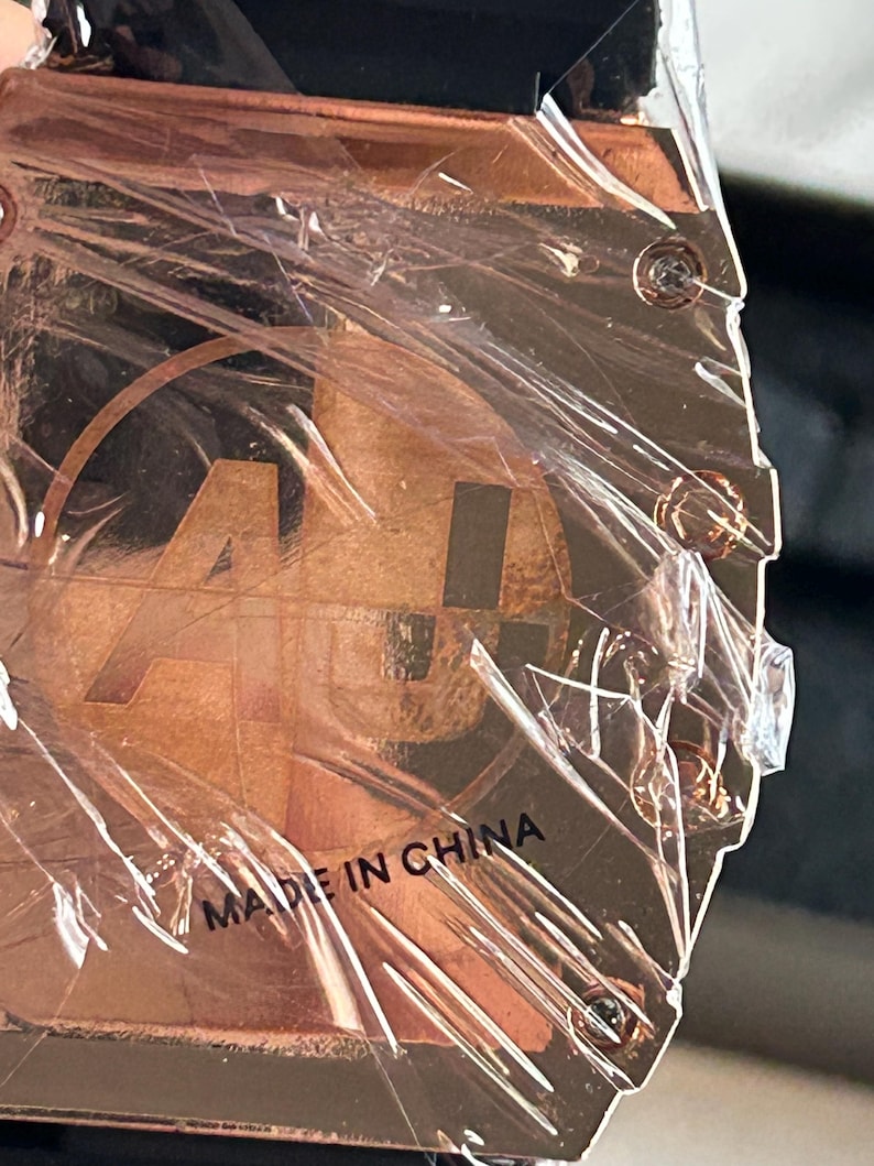 May include: A square-shaped copper-toned watch case back featuring a circular logo with the letters AU inside. The metal surface is covered in a protective clear plastic film and includes four visible screws at the corners with the text MADE IN CHINA.