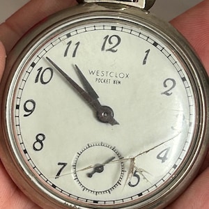 May include: A silver pocket watch with a white face and black numbers. The watch has a cracked glass face and the text "Westclox Pocket Ben" is printed on the face.