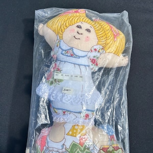 May include: A Cabbage Patch Kids doll in a plastic bag. The doll has blonde hair with pink ribbons, a blue dress with floral accents, and the text "Hi! My name is Amy Sue". The doll's packaging includes the Cabbage Patch Kids logo.