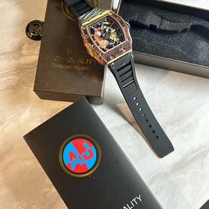 May include: A wristwatch with a black silicone strap and a rose gold-toned tonneau-shaped case featuring a tiger graphic on the dial. The watch is presented with two black boxes, one displaying the text HIGH-QUALITY Designed in USA and an AJ logo.