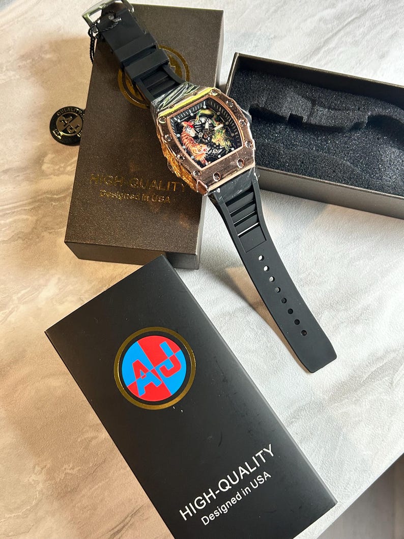 May include: A wristwatch with a black silicone strap and a rose gold-toned tonneau-shaped case featuring a tiger graphic on the dial. The watch is presented with two black boxes, one displaying the text HIGH-QUALITY Designed in USA and an AJ logo.