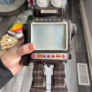 May include: A vintage brown toy robot with a rectangular screen and circular eye-like details. The robot has red accents on its feet and a row of red and yellow lights. A retro-themed collectible.